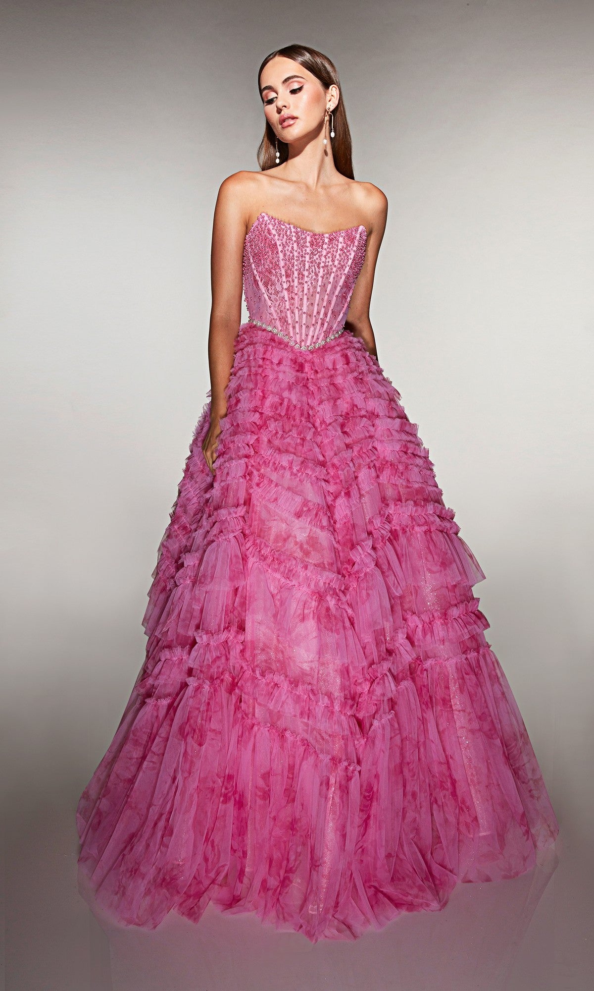 A woman poses against a plain background in the Alyce 61981 Long Prom Dress, a pink strapless ball gown with a beaded corset bodice and a voluminous ruffled tulle skirt.